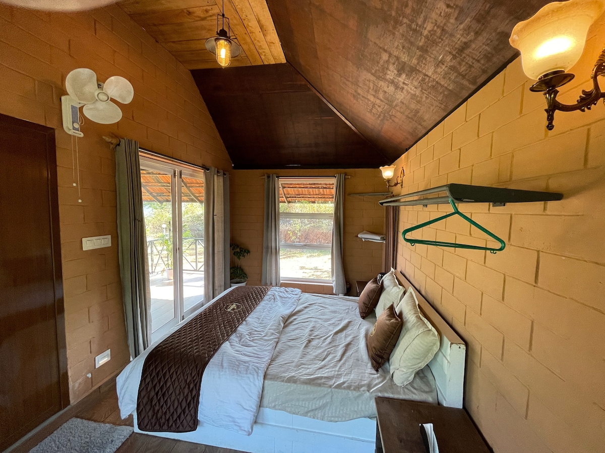 A cozy bedroom features a spacious bed with a light-colored blanket and several decorative pillows. Large windows provide natural light and a view of the outdoor area. A fan hangs from the ceiling, and soft lighting is provided by wall sconces.