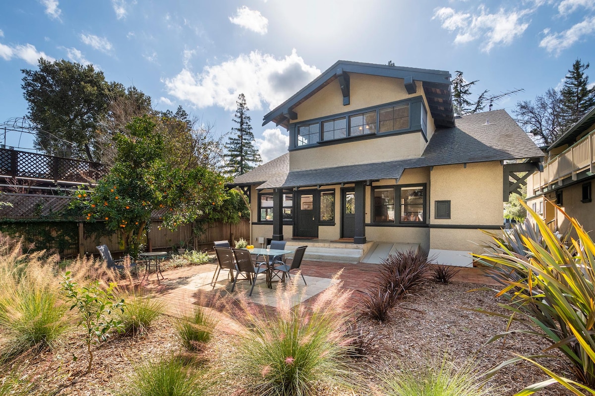 The exterior of a spacious Craftsman home is displayed, featuring a well-maintained backyard with a patio area furnished with a table and chairs. Lush greenery, including ornamental grasses and small trees, enhances the outdoor space. The home's architectural details, including a sloped roof and large windows, are visible.