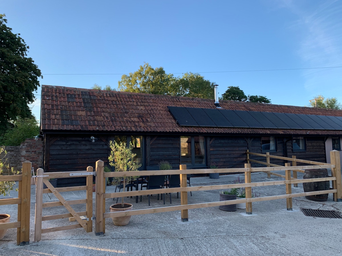 A charming barn-style building with a rustic wooden exterior is featured, complemented by solar panels on the roof. A fenced area is visible in the foreground, with outdoor seating arrangements and potted plants enhancing the entrance.