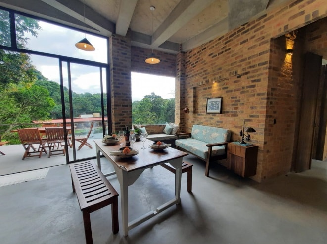 An open living space features a wooden dining table set for meals, complemented by a comfortable sofa near large windows that offer views of lush greenery. The room showcases exposed brick walls and minimalistic furnishings, creating a warm and inviting atmosphere.