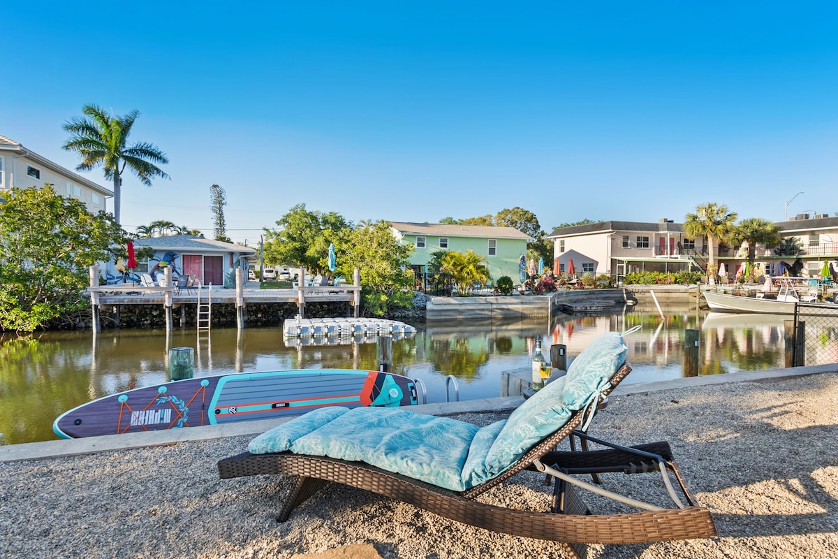 A comfortable lounge chair is positioned on the sandy shore, overlooking a calm canal. Lush greenery is present along the water's edge, and various boats are moored nearby, showcasing the area's access to water activities. A clear blue sky provides a serene backdrop.