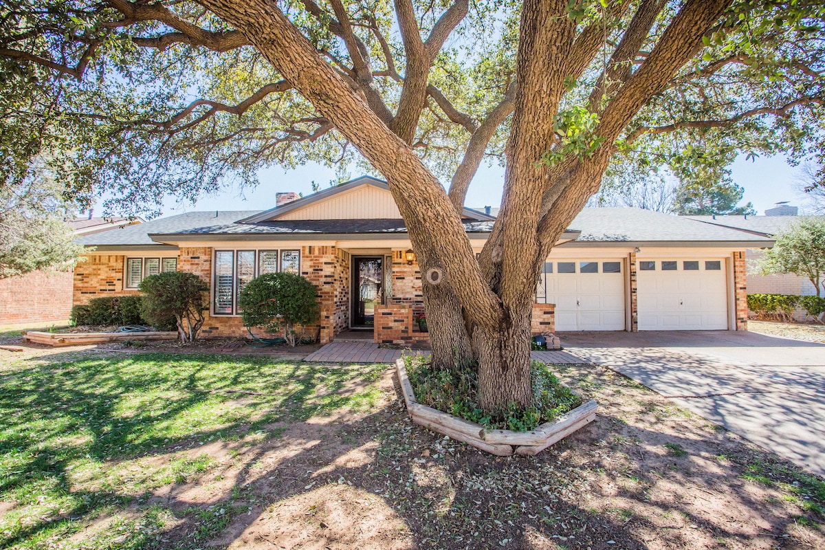 The exterior of the home is framed by a large tree, providing shade over the manicured lawn. The brick facade features multiple windows, and a spacious driveway leads to a two-car garage. Neatly landscaped bushes complement the inviting entrance.