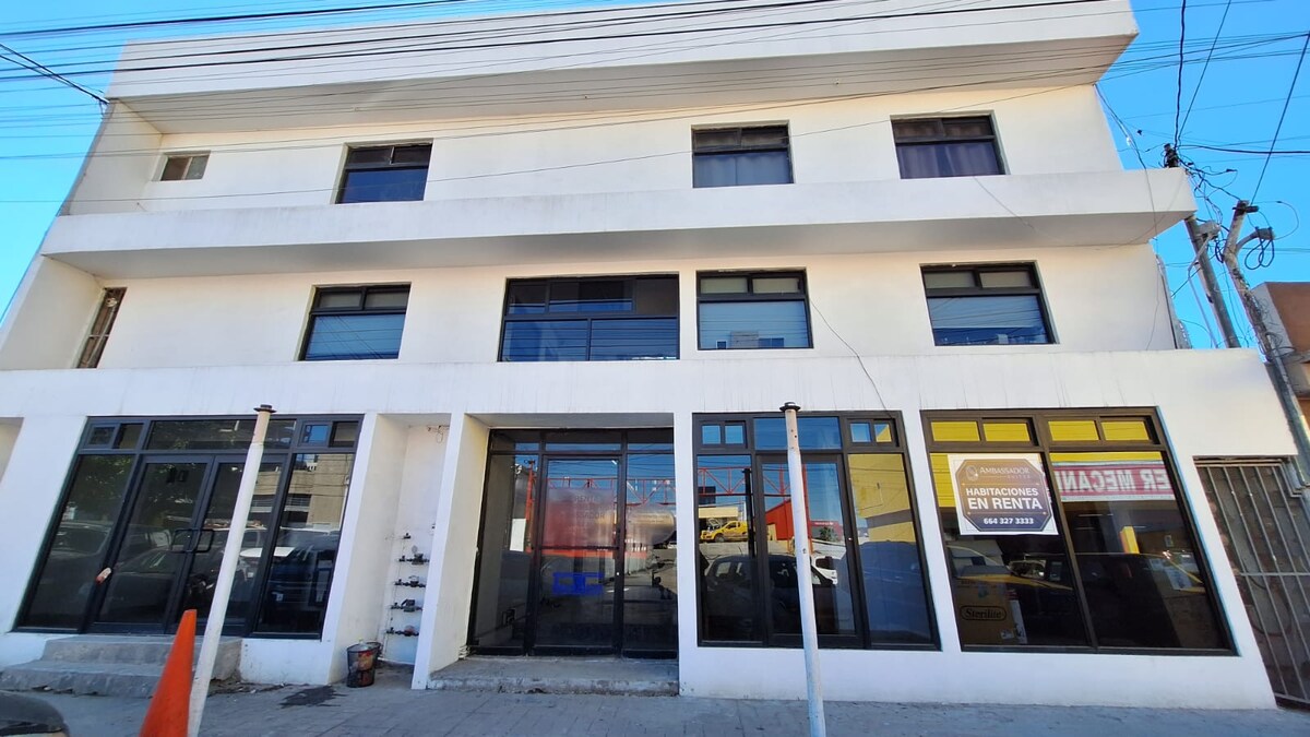 The exterior of a three-story building features a modern design with large windows that allow natural light. A variety of retail spaces are visible at street level, framed by a clean, minimalist facade. The area is well-lit, and nearby infrastructure is noticeable.