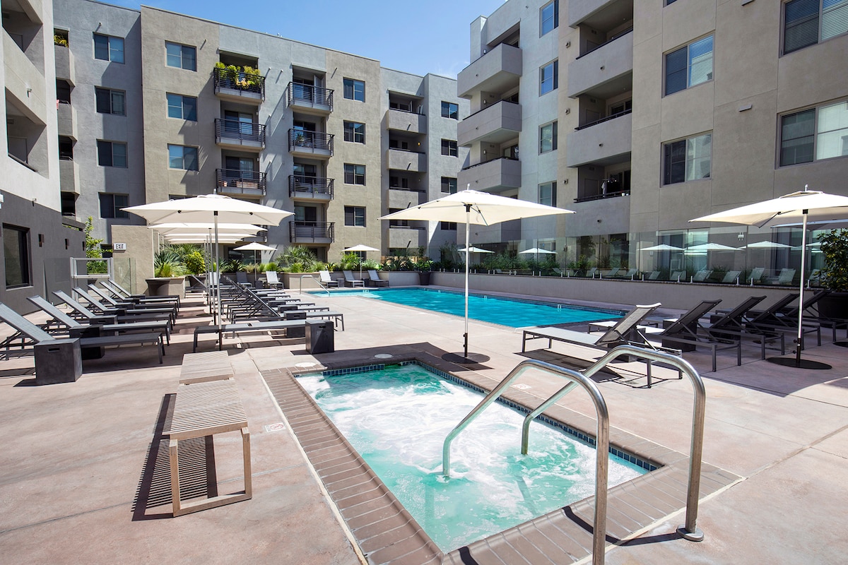 A shared outdoor pool area is depicted, featuring a large swimming pool complemented by lounge chairs under large umbrellas. An adjacent hot tub is visible, surrounded by a textured concrete surface, with nearby greenery adding to the setting.