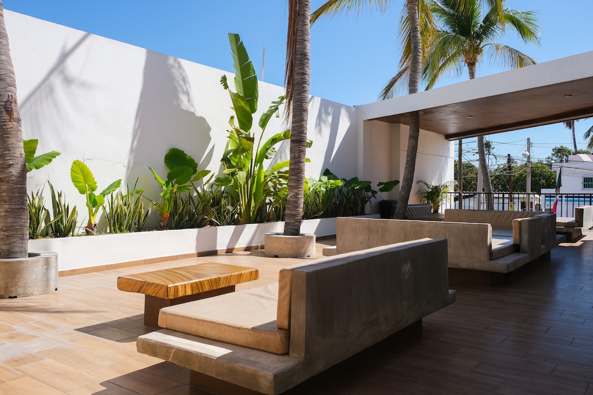 An outdoor lounge area is presented, featuring modern concrete seating arranged around a wooden table. Lush greenery, including palm and banana plants, adds natural beauty to the space. Sunlight filters through, creating a bright and inviting atmosphere.