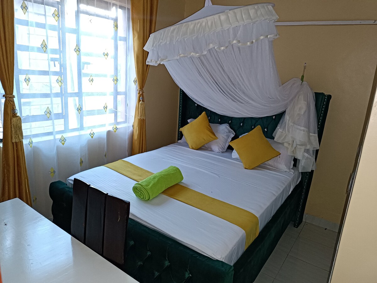 A well-furnished bedroom features a spacious bed with a white canopy draped above. Decorative yellow cushions are placed on the bed, while a bright green towel is arranged neatly. Soft natural light filters through the patterned curtains, illuminating the inviting space.