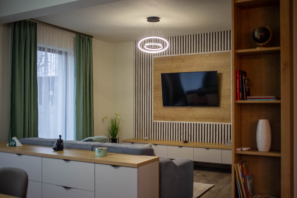 A modern living area is highlighted by a wall-mounted television centered above a light wood feature. Soft grey sofas are positioned around a light-colored sideboard, while natural light flows through sheer drapes, creating a relaxed atmosphere.