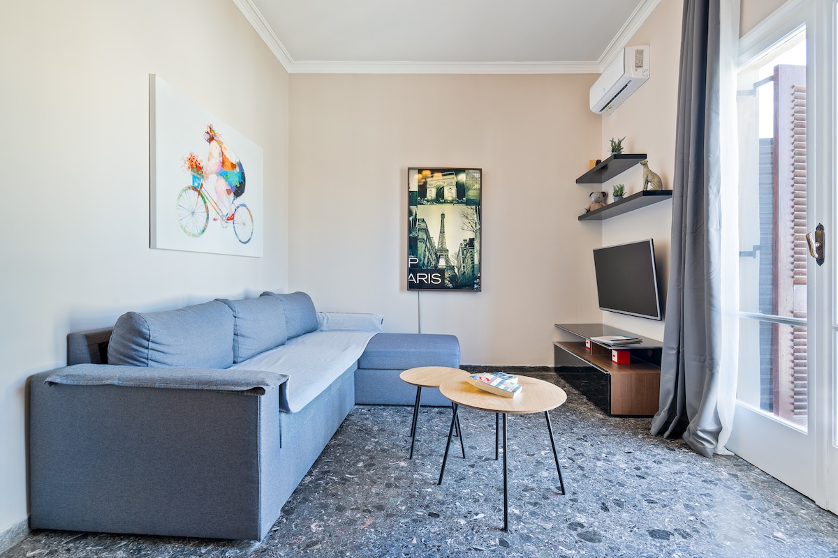 A spacious living room features a large gray sectional sofa and a natural wood coffee table. A flat-screen television is mounted on the wall, accompanied by shelves displaying decor items. Light enters through a glass door leading to a small balcony, enhancing the bright atmosphere.