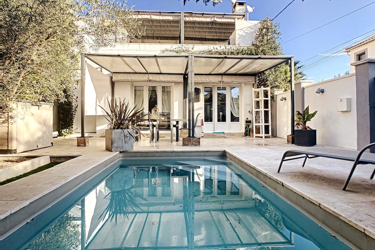 A private outdoor area features a sleek swimming pool surrounded by a stone deck. Dining and lounge furniture are arranged under a modern pergola, with greenery and decorative plants adding a touch of nature. Sun loungers are positioned for relaxation.
