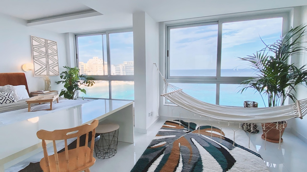 A bright living space is furnished with a spacious bed and a cozy hammock. Large windows offer expansive views of the sea, while a decorative area rug adds color. Potted plants enhance the airy atmosphere, and a dining table provides additional functionality.