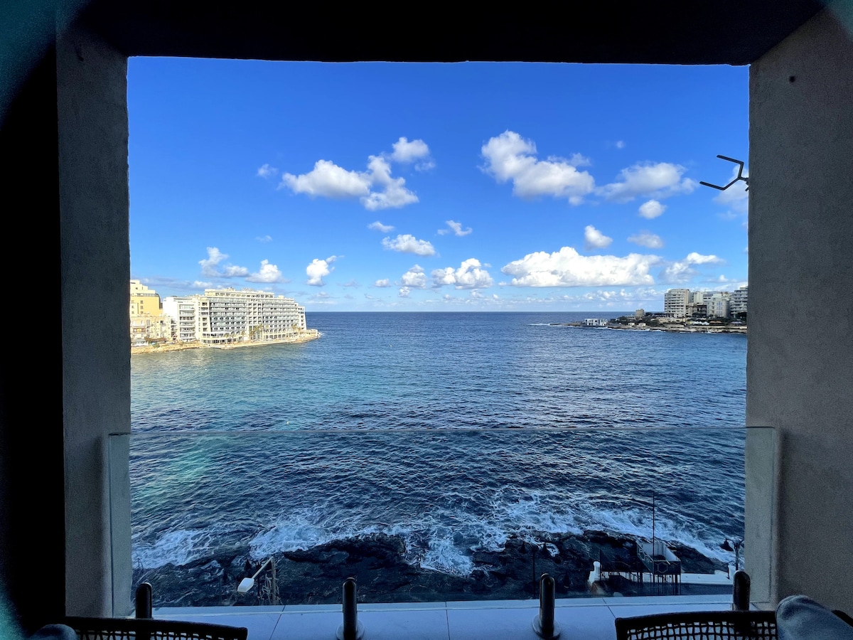 A panoramic view of the sea is captured through a large opening, showcasing calm waters meeting a clear blue sky dotted with white clouds. The coastline features buildings along the shore, while rocky outcrops are visible in the foreground.