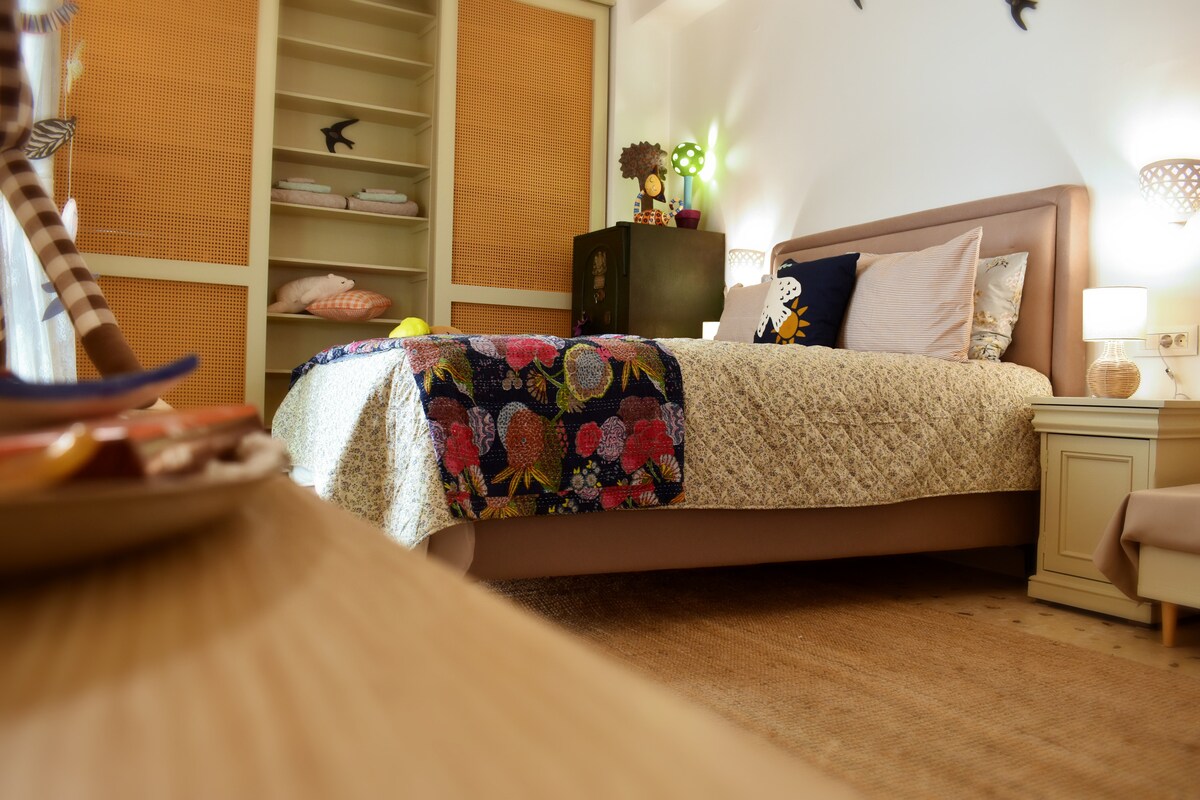 A cozy sleeping area is highlighted by a quality double bed with decorative pillows. A colorful bedspread featuring floral patterns covers the bed. Warm lighting is provided by bedside lamps, and a large shelving unit is visible in the background.