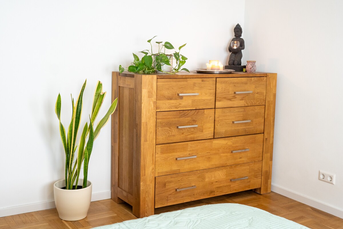 A wooden dresser with six drawers occupies one corner of the room. On top, potted plants and a small statue are displayed, while a plant in a beige pot adds freshness to the space. The walls are painted white, complementing the natural wood tones.