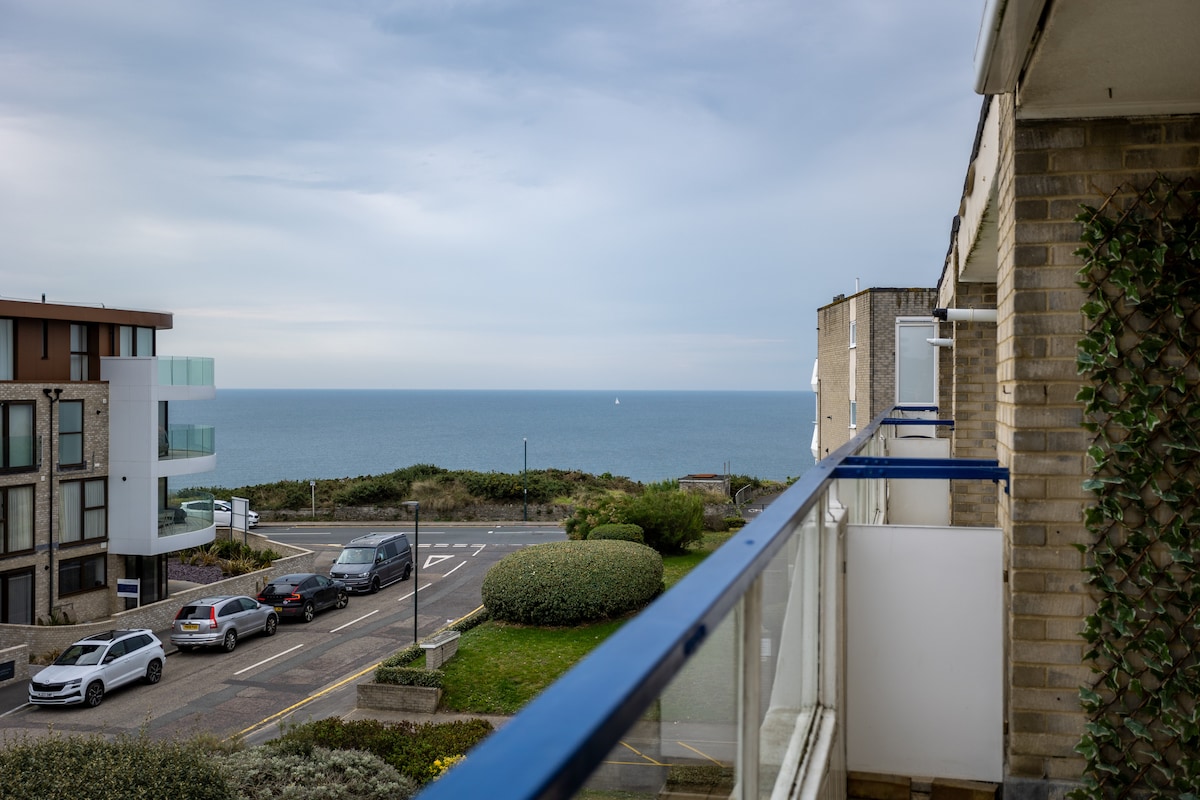A view from the balcony showcases the expansive sea, extending towards the horizon. The coastline is lined with greenery, framed by the apartment's railing. A quiet street is visible below, with buildings positioned on either side, creating a serene atmosphere.