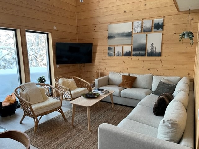 A inviting living area features large windows that allow natural light to brighten the space. A comfortable sectional sofa and two armchairs provide ample seating. A flat-screen television hangs on the wall, while a coffee table is adorned with books and decor. Natural wooden walls enhance the cozy ambiance.