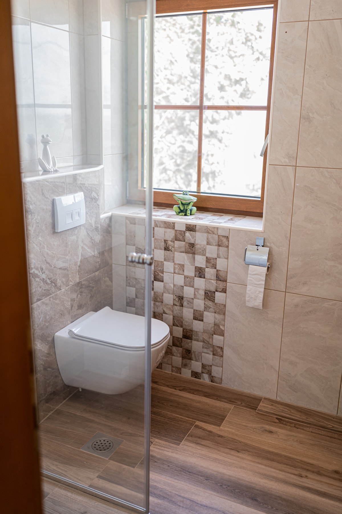 A modern bathroom features a glass-enclosed shower and a white toilet. The walls are adorned with elegant tiles, complemented by a small potted plant on the window sill, allowing natural light to fill the space.