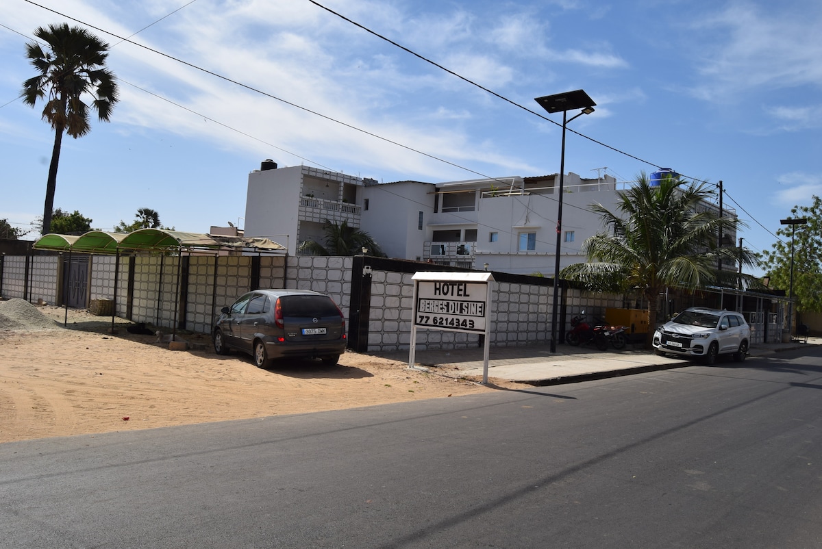 A modern hotel building is displayed against a clear blue sky, with a sandy area in the foreground. The entrance is marked by a prominent sign, and several palm trees frame the property. Parking spaces are visible along the road, highlighting easy accessibility.