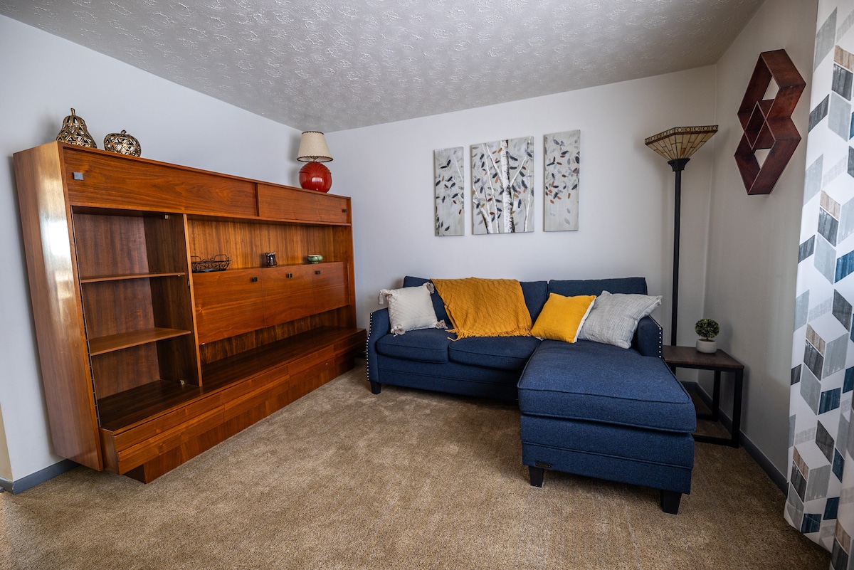 A cozy living area features a blue sectional sofa adorned with throw pillows and a mustard-yellow blanket. A wooden bookshelf stands against the wall, while three framed artworks add visual interest. A patterned lamp and a geometric decor piece complete the inviting space.