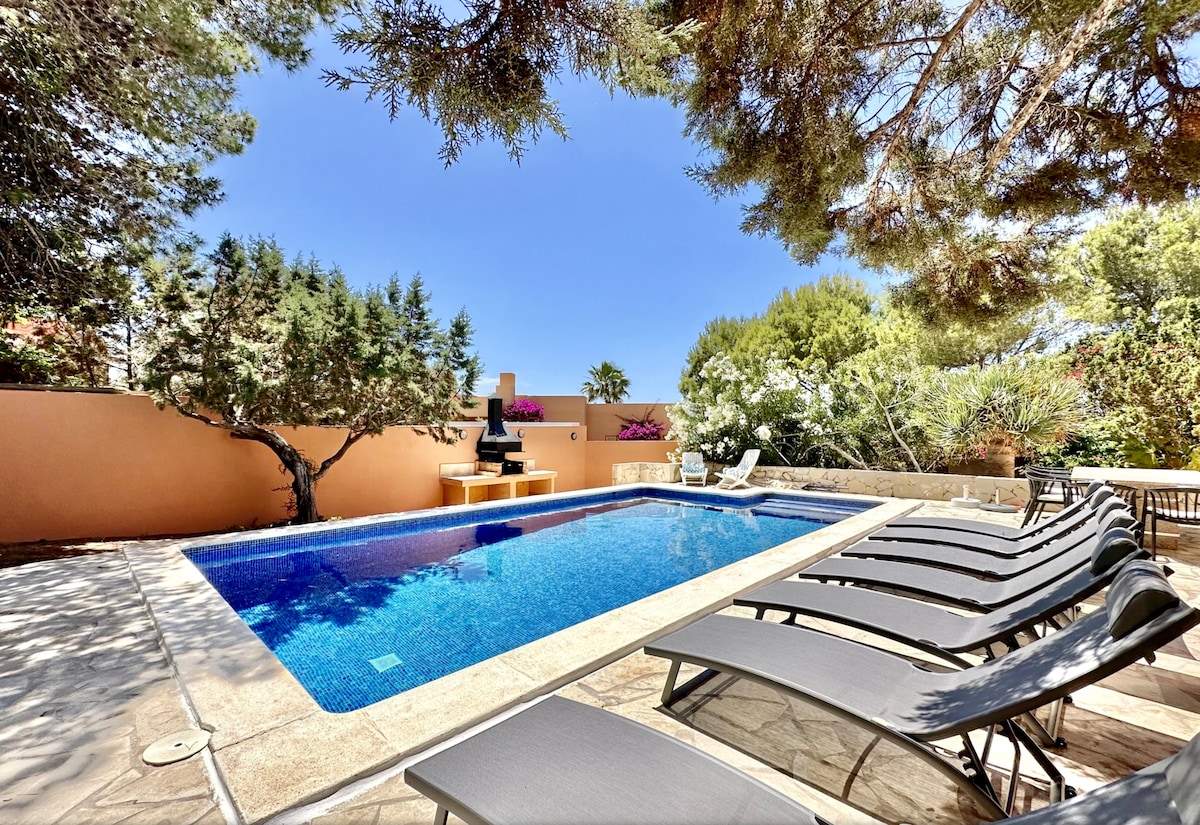 An outdoor swimming pool is surrounded by comfortable lounge chairs. Lush greenery and flowering shrubs provide a serene backdrop. A small seating area with a water feature is positioned near the pool, enhancing the tranquil environment for relaxation.
