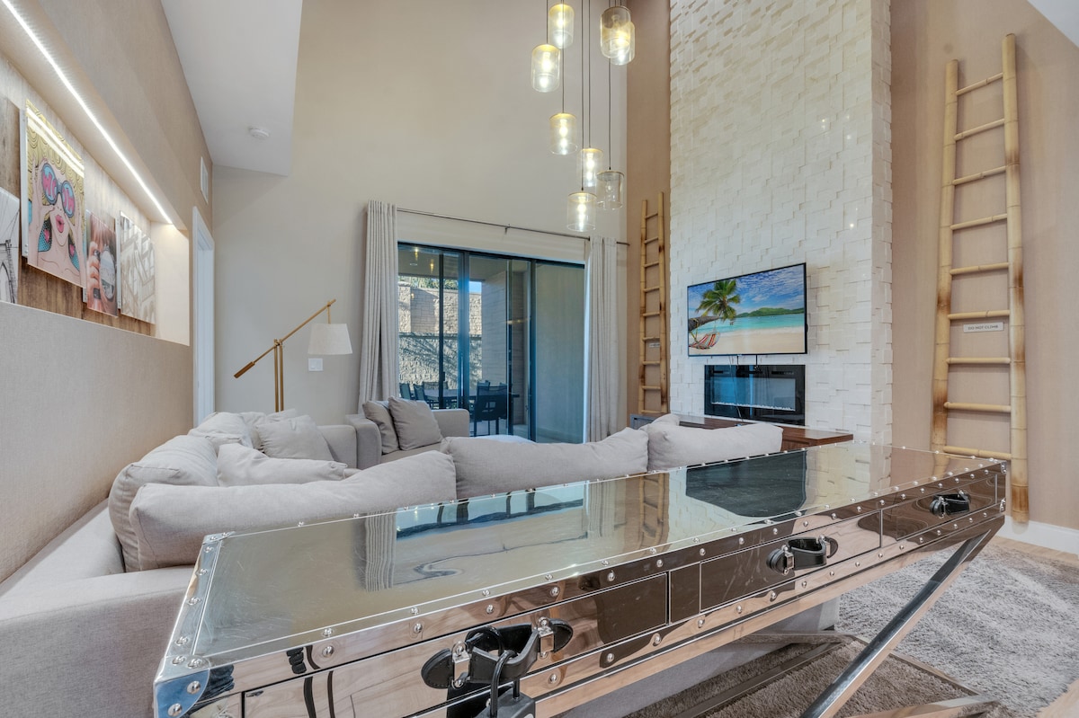 The living area features a spacious sectional sofa arranged around a glass-top coffee table. A mounted television is positioned above a modern fireplace, with artwork displayed on the walls. Natural light streams in through sliding glass doors leading to an outdoor space.