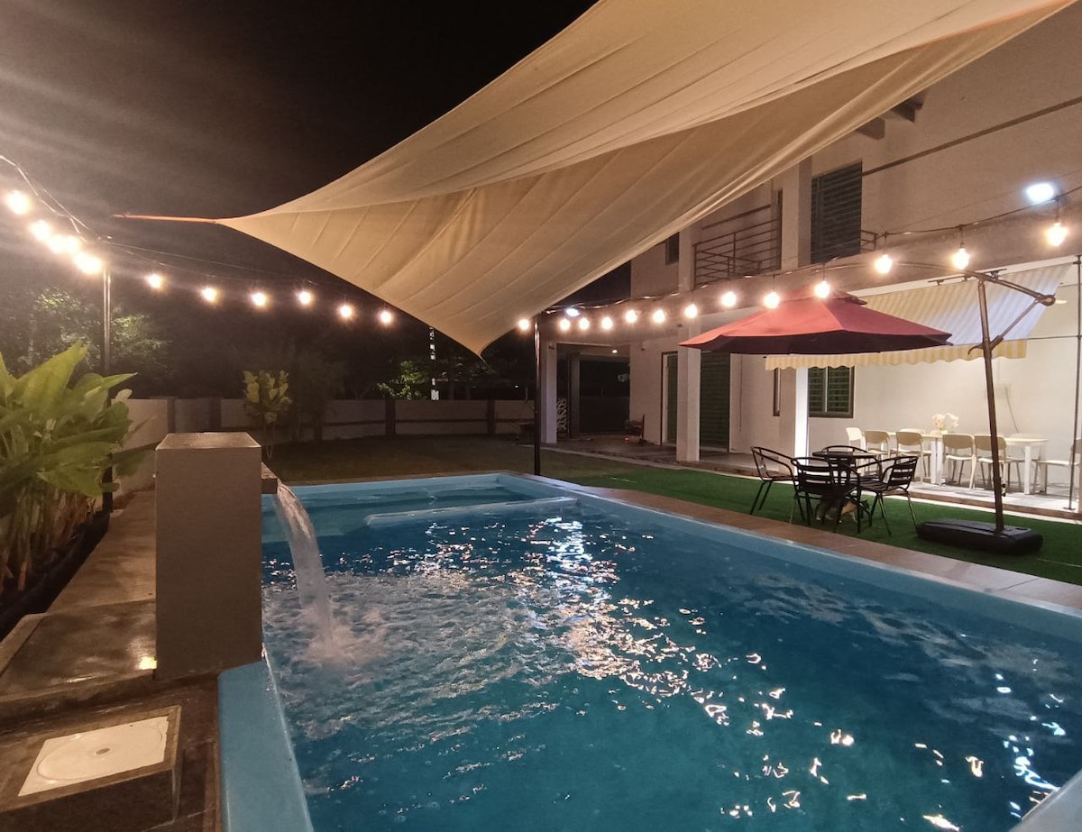 An outdoor area features a pool illuminated by soft lighting. A large sail provides shade, while string lights add ambiance. Various seating options, including tables and chairs, are arranged nearby, surrounded by lush greenery and a tastefully designed exterior.