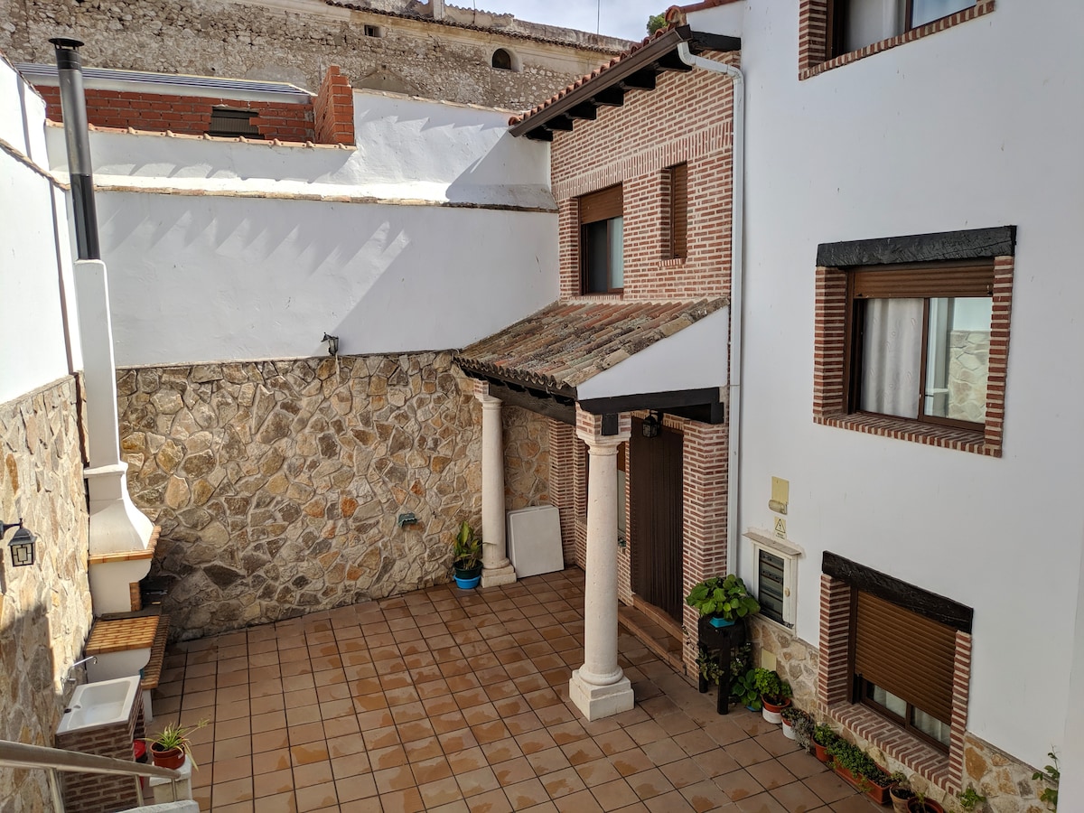 A charming courtyard is framed by stone walls and complemented by terracotta tiles. Pillars support a small overhang, creating a shaded area. Potted plants add touches of greenery alongside windows, while natural light illuminates the space, contributing to a sense of openness.