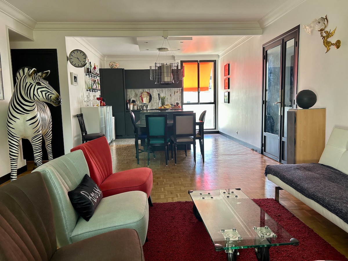 A spacious living area features a variety of seating options, including a colorful sofa and an accent chair. A sleek glass coffee table sits on a textured rug. In the background, a dining space is visible, with vibrant artwork enhancing the room's character.