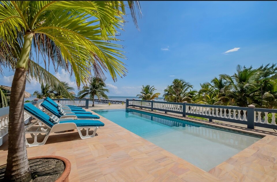 A serene infinity pool is surrounded by comfortable lounge chairs, inviting relaxation under the clear blue sky. Lush palm trees frame the view, while the calm waters of the sea are visible in the background, enhancing the tranquil outdoor space.