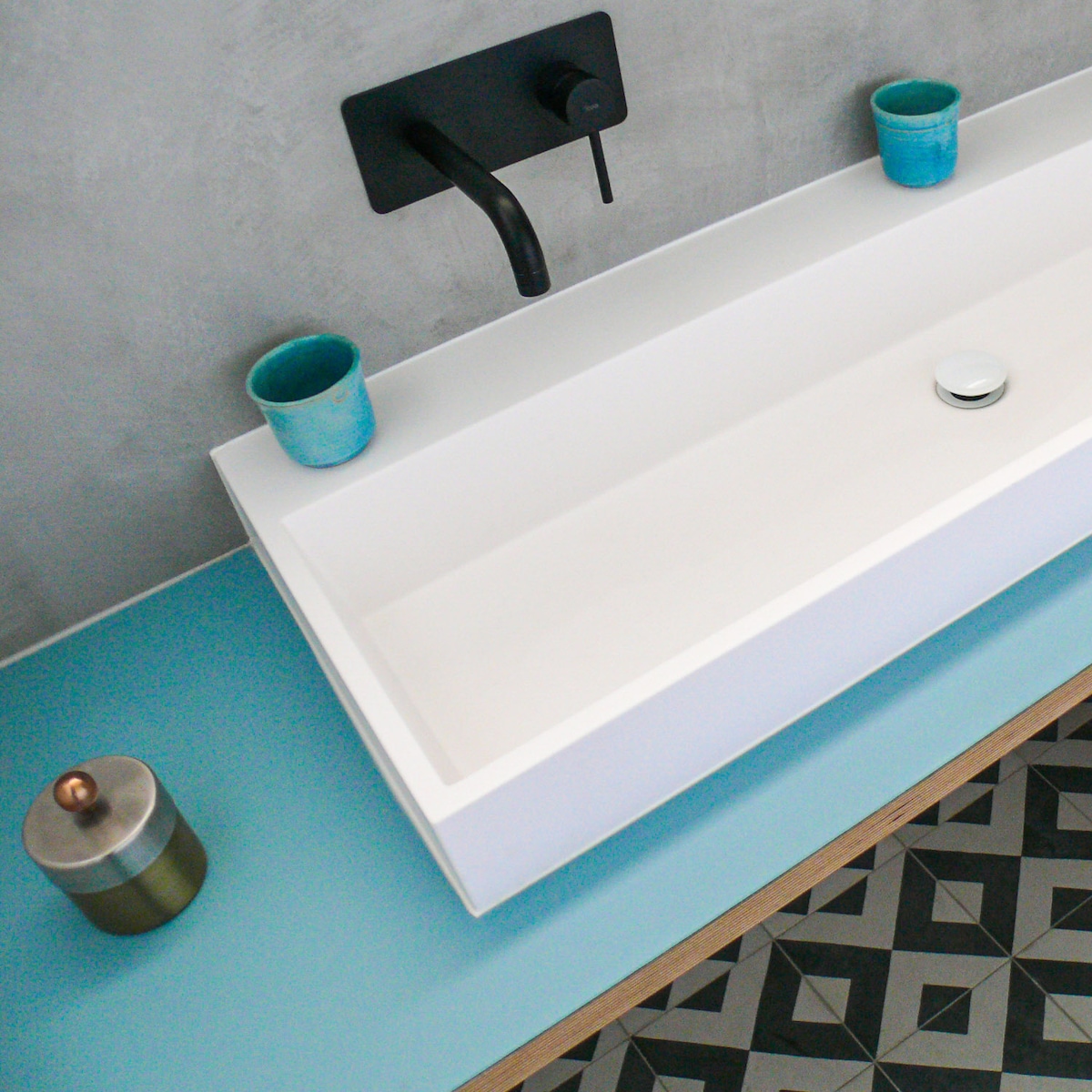The bathroom features a minimalist design with a white, rectangular sink positioned on a seafoam green countertop. Two soft blue cups are placed at either end of the sink, complemented by a black faucet above. The floor showcases a geometric tiled pattern in monochrome tones.