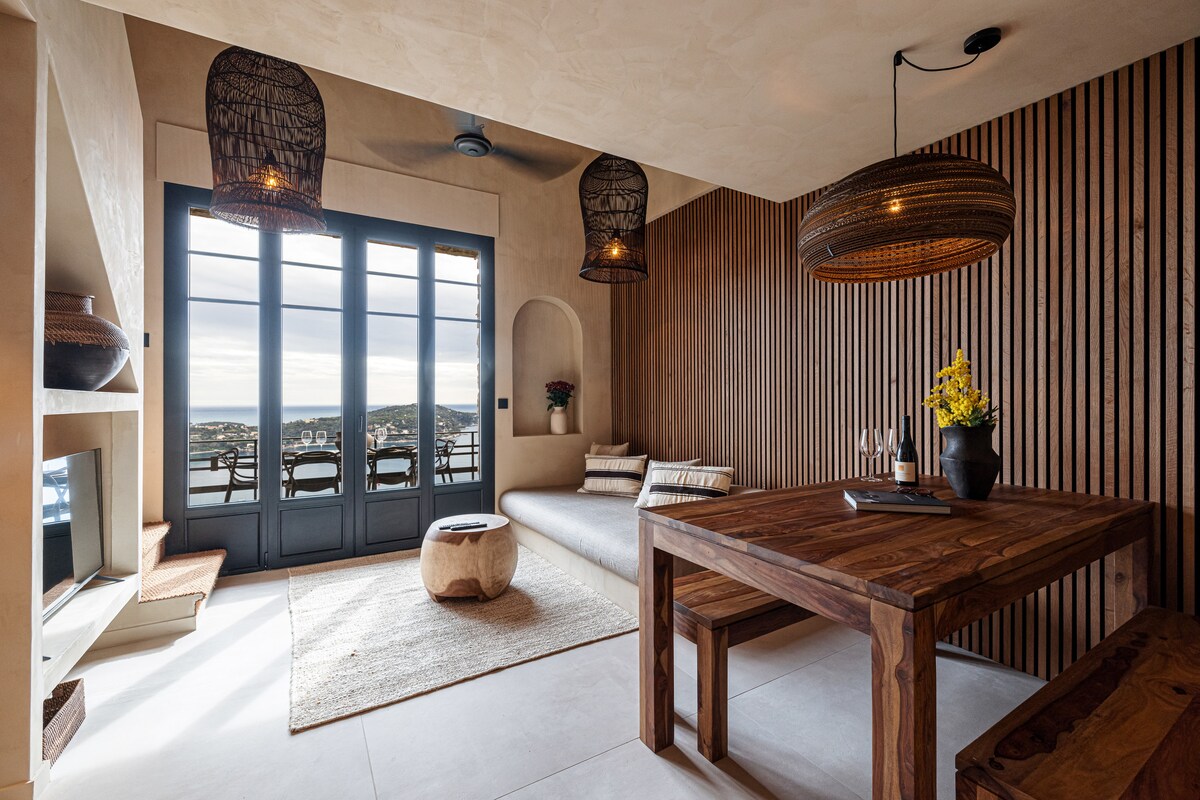 A spacious living area features a wooden dining table and a cozy seating nook, all enhanced by natural lighting from the large glass doors. Textured walls and artistic lighting fixtures contribute to a warm yet modern ambiance, with a view of the bay visible through the doors.