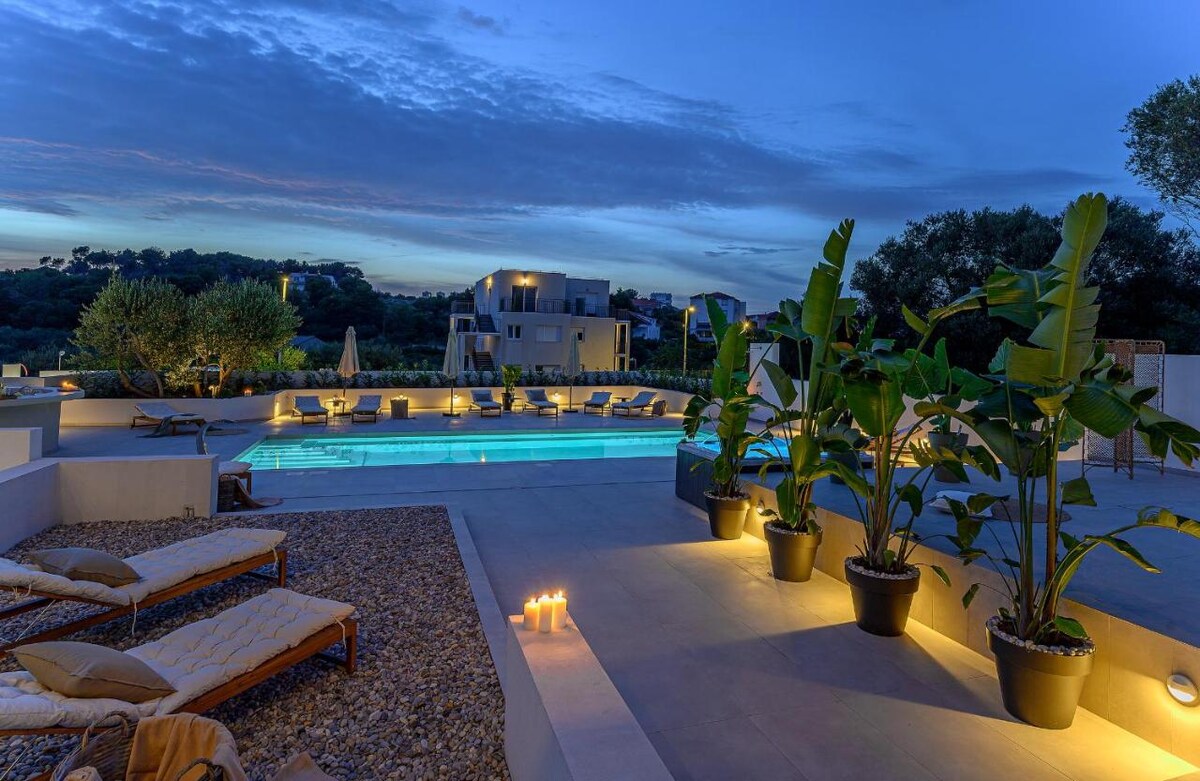 An outdoor area features a pool illuminated softly at dusk, surrounded by loungers and vibrant green plants. The space is enhanced by candles placed on a gravel surface, creating a serene retreat atmosphere for relaxation.