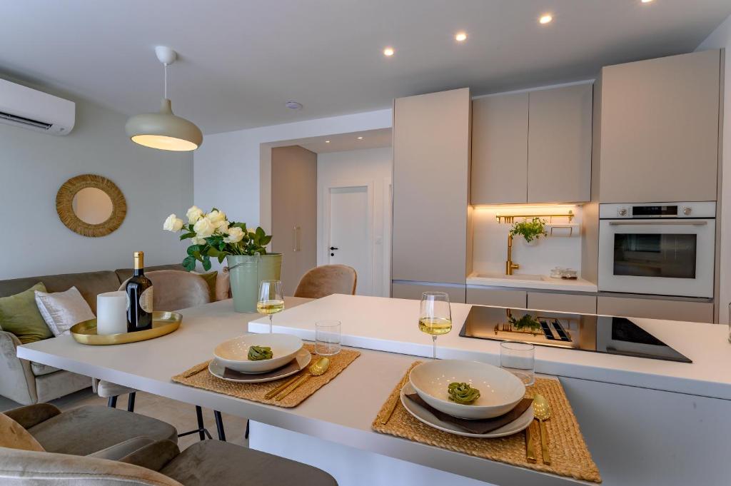 A modern kitchen is showcased, featuring sleek cabinetry and a central island. Two place settings are elegantly arranged with glassware and plates, complemented by a vase of white flowers. Soft lighting enhances the spacious atmosphere, while a focus on functionality is evident throughout.