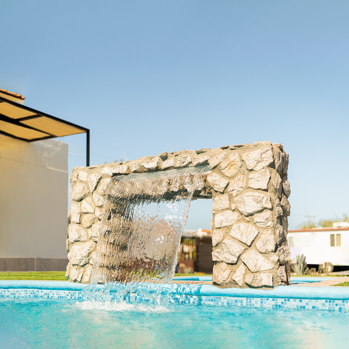 A stone feature creates a gentle waterfall that flows into the clear blue pool, enhancing the outdoor space. The backdrop includes a structure with a shaded area, inviting relaxation and enjoyment of the serene surroundings.