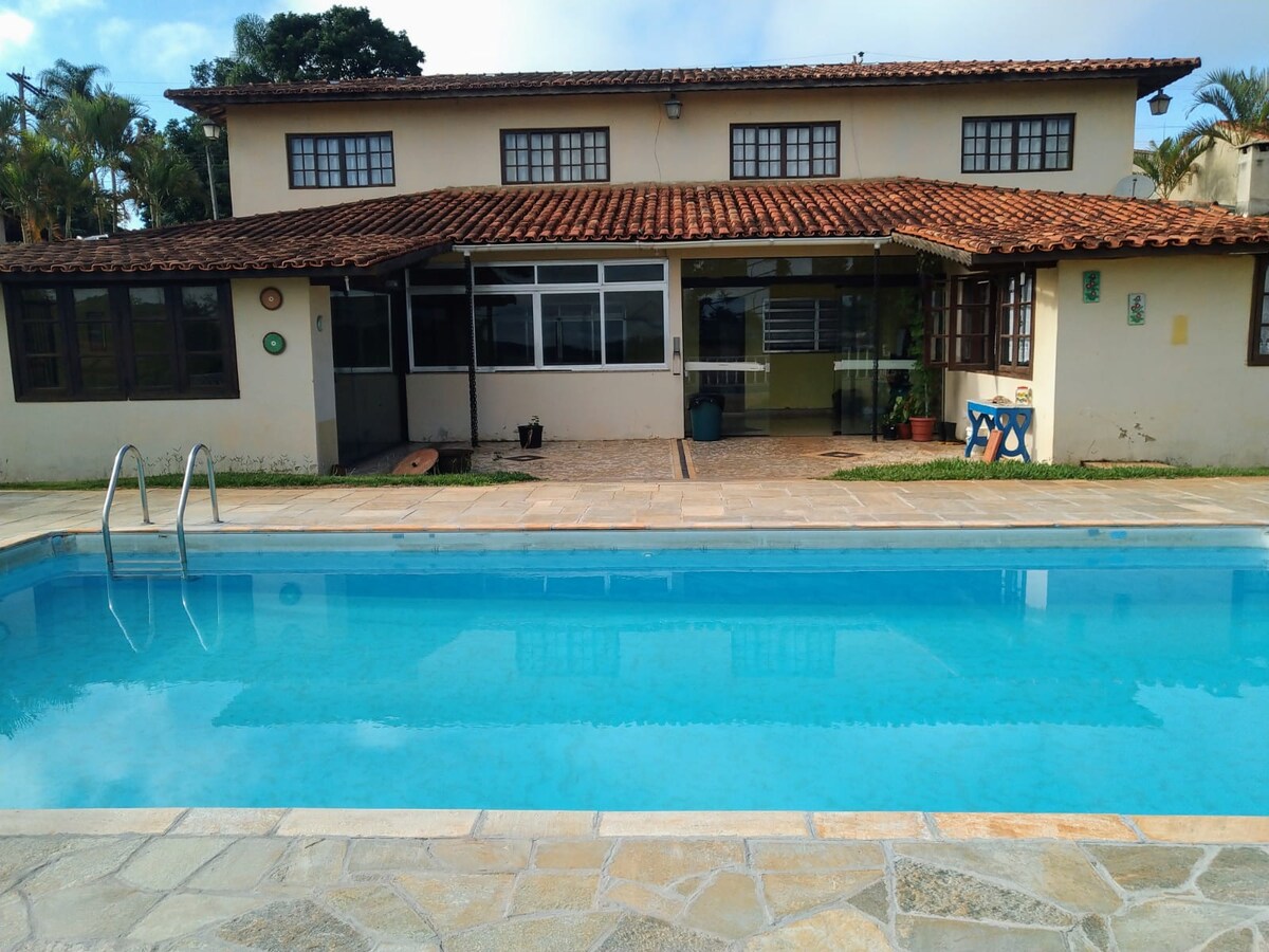 A spacious outdoor area is presented, featuring a clear swimming pool framed by light-colored stone tiles. The two-story house with a tiled roof is visible in the background, showcasing large windows and an outdoor seating area. Lush greenery adds to the inviting atmosphere.