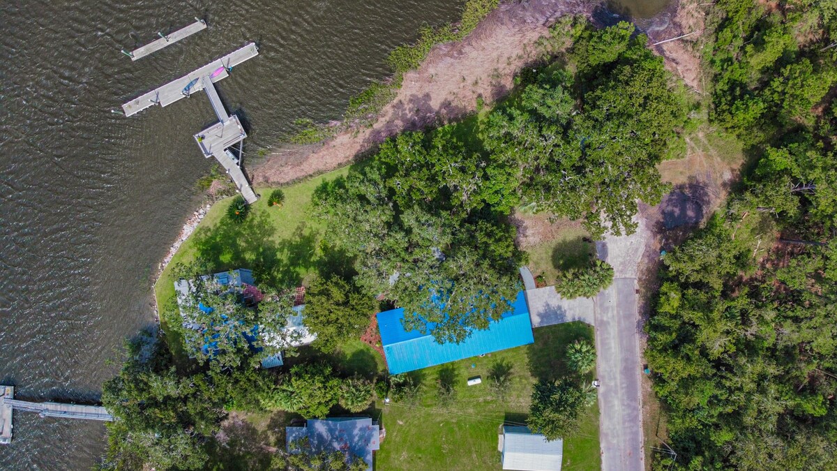 An aerial view reveals a spacious property bordered by water, featuring a private dock for boating activities. Green vegetation surrounds the landscape, with a blue-roofed building and other structures visible. The serene setting offers a blend of natural beauty and accessibility.