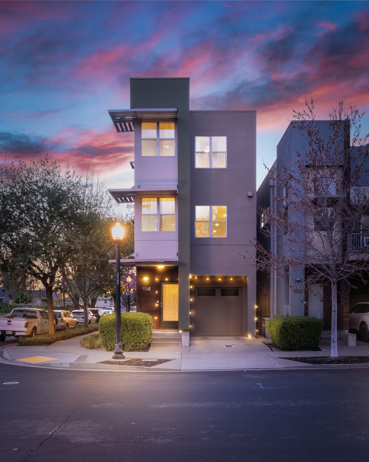 The tri-level home is featured at dusk, with modern architecture and large windows. Soft ambient lighting outlines the entryway and balcony. Surrounding greenery and vehicles are visible, all complemented by a colorful sunset sky.