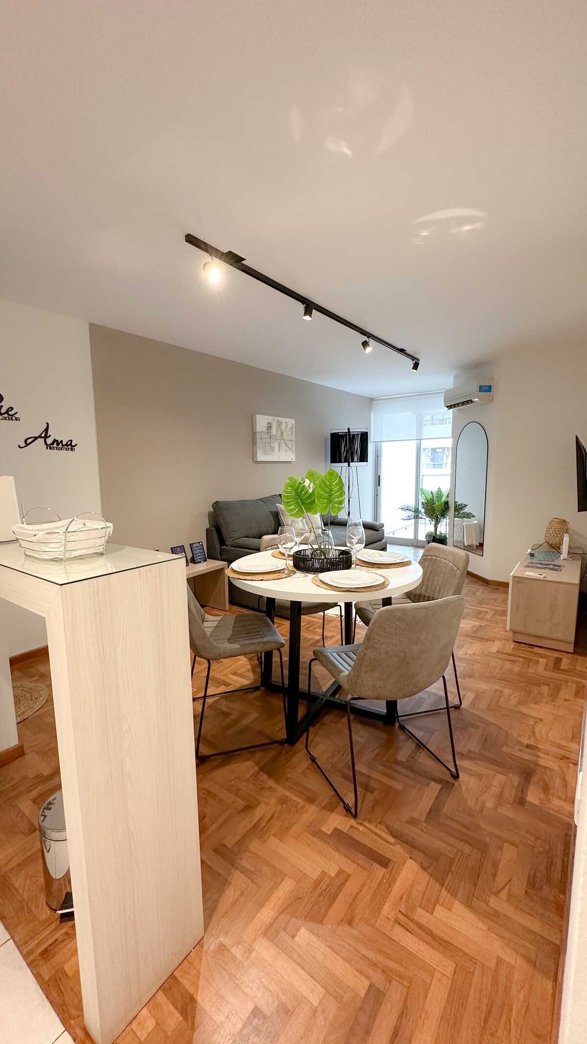 A dining area features a round table set with dishware and decorative items. Four grey chairs surround the table, and a sofa is positioned nearby. Large windows allow natural light to brighten the space, with plants adding a touch of greenery.