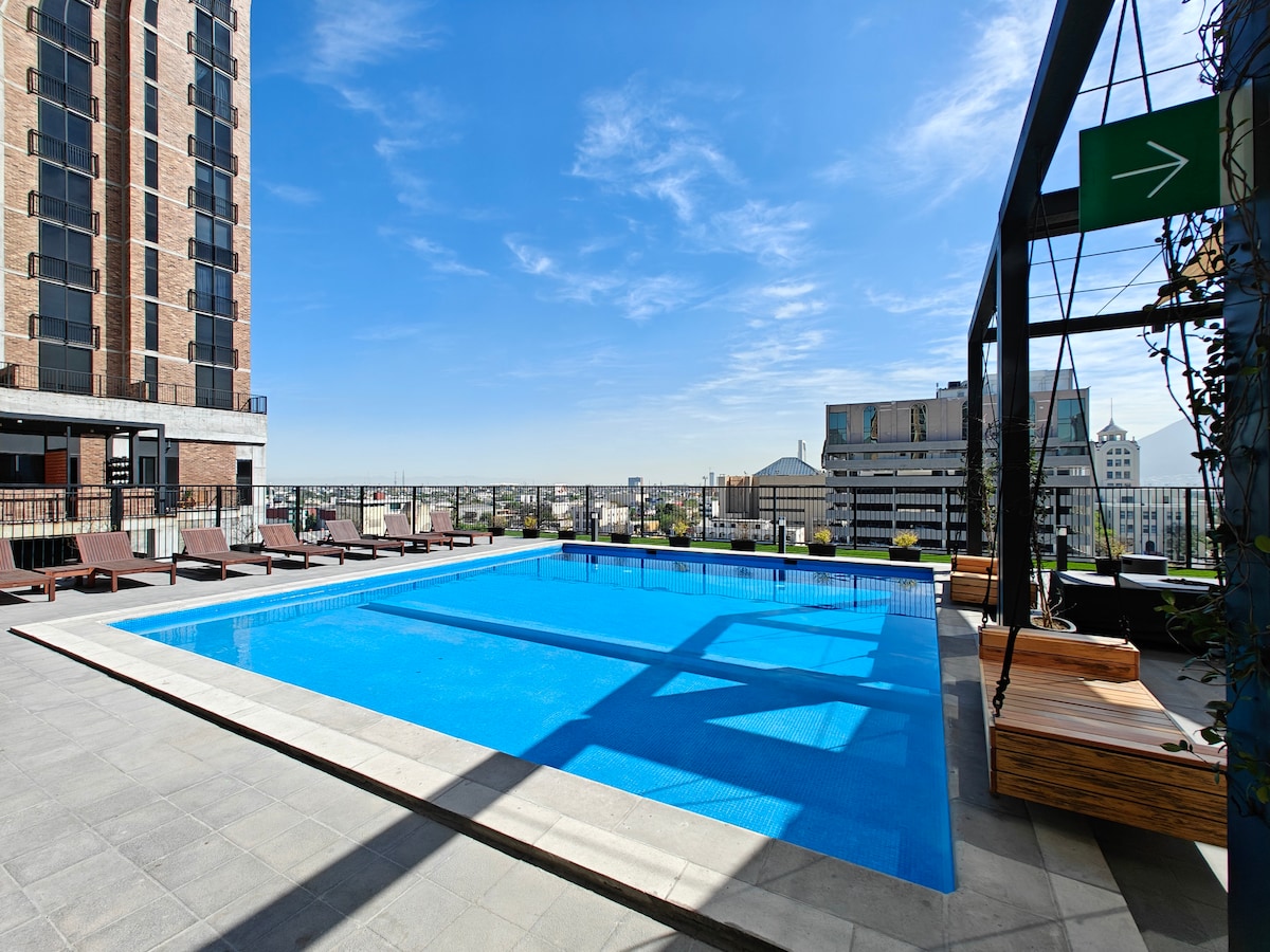 A rooftop pool area features a large, clear swimming pool surrounded by lounging chairs and shaded seating. The urban skyline is visible in the backdrop under a bright blue sky, creating a refreshing outdoor space for relaxation.