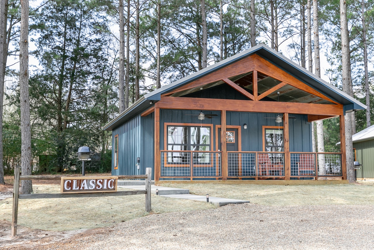 A cabin with a blue exterior features a large front porch and wooden accents. Large windows provide views of the surrounding pine trees. The word 'CLASSIC' is displayed prominently in front of the cabin, adding to its charm. A grill is positioned nearby.