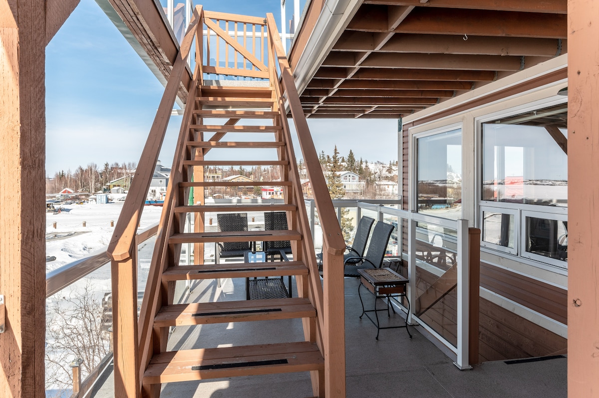 A wooden staircase leads to an upper-level balcony, framed by wooden railings. The balcony is furnished with black outdoor chairs and a small table, overlooking scenic views of the surroundings. Large windows provide natural light to the adjacent area.