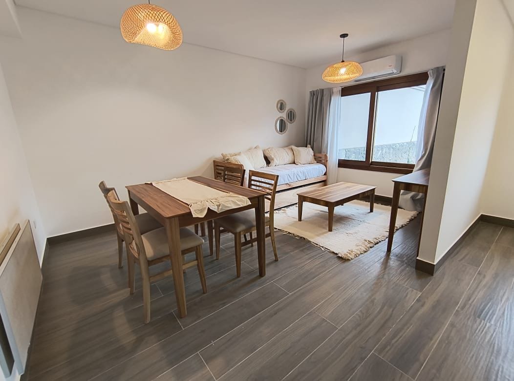 A modern living space features a light-colored sofa, a wooden dining table with chairs, and a coffee table on a woven rug. Natural light enters through a large window, and decorative elements include wall mirrors and pendant lights.