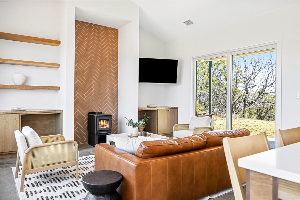 A modern living area showcases a leather sofa and two light armchairs, positioned near a natural wood shelving unit. A compact fireplace is set against a textured wall, while large glass doors provide a view of the outdoors, enhancing the inviting ambiance.