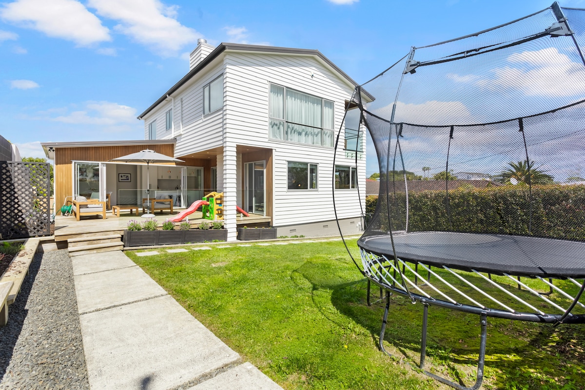 The exterior of the home features a spacious backyard with a large trampoline and children's climbing frame. A patio area provides outdoor seating, and a sliding door opens to the living space. Lush greenery surrounds the yard, enhancing the overall landscape.