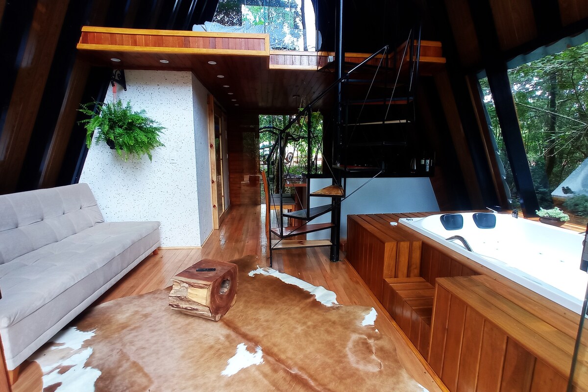 An open living space features wooden flooring and a spiral staircase leading to a loft area. A soft sofa faces a natural wood accent table, while a jetted tub is incorporated into the design. Green foliage is visible through large windows, connecting the indoors to nature.