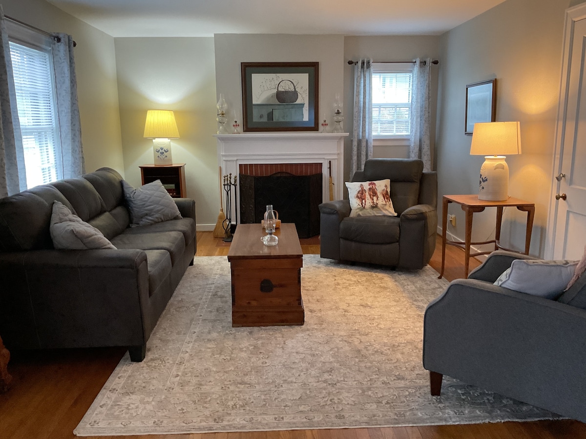 A comfortable living room is featured, showcasing a large area rug beneath a wooden coffee table. Two sofas and an armchair are arranged around the space. Soft lighting is provided by table lamps, and a fireplace is set against a neutral wall, complementing the décor.