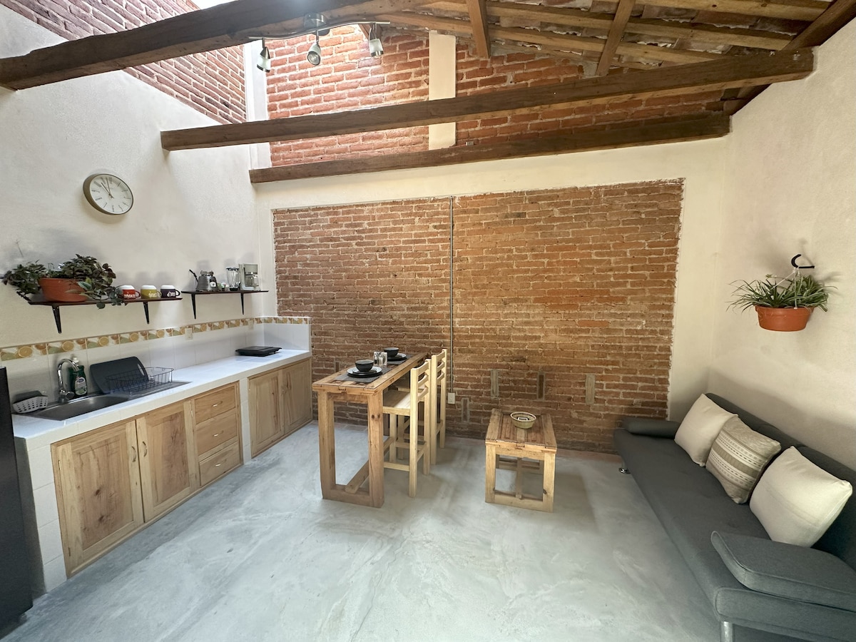 A modern kitchen area features exposed brick walls and wooden beams overhead. A wooden table is positioned in the center, with three chairs nearby. Appliances are neatly arranged on the countertop, complemented by potted plants and natural light entering through a window.