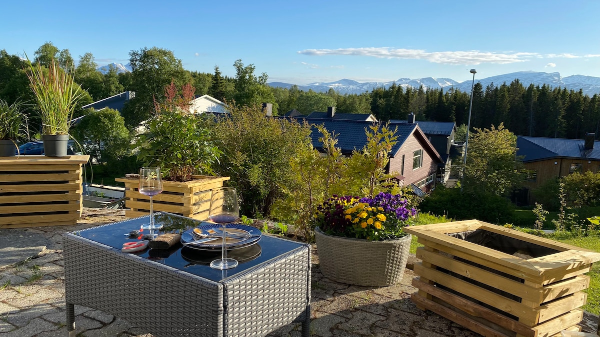An outdoor seating area is presented with a glass-top table surrounded by potted plants and colorful flowers. Scenic views of distant mountains and greenery are visible in the background, offering a serene outdoor retreat.