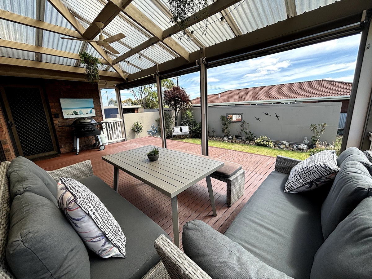 A covered outdoor deck area features a sleek table at the center, surrounded by comfortable gray seating with decorative pillows. A barbecue grill is positioned nearby, and a grassy area with low maintenance landscaping is visible beyond the deck.