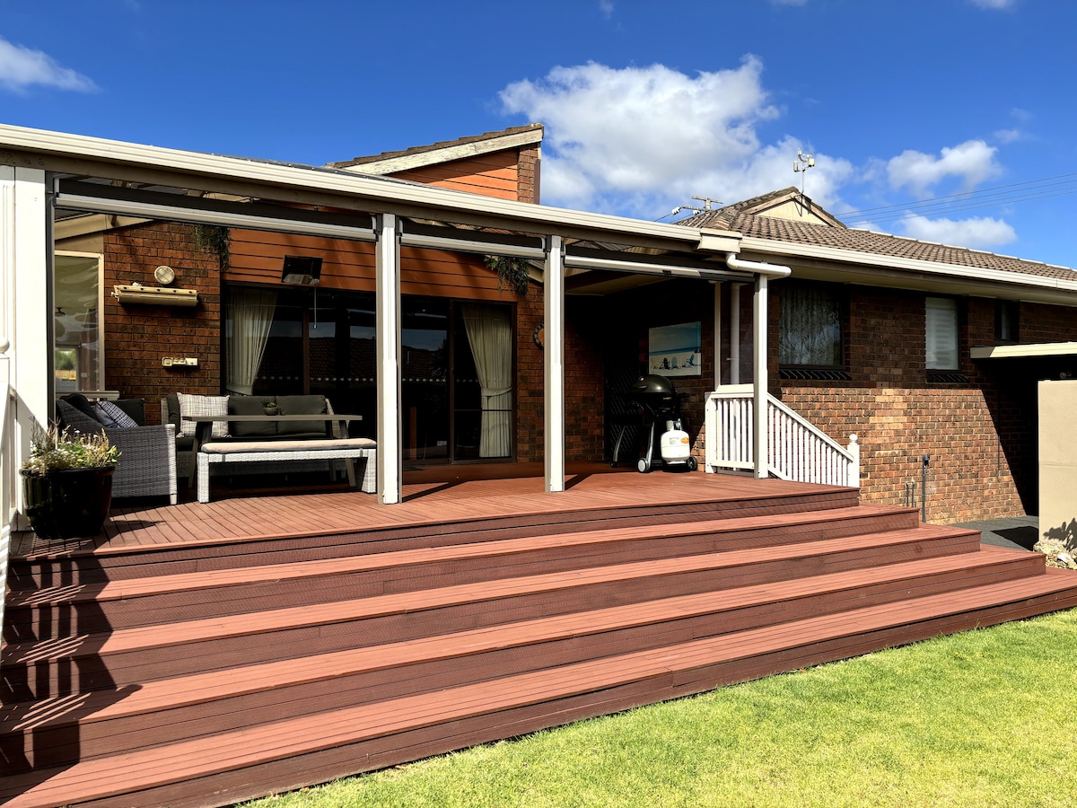 The outdoor deck showcases a spacious area for relaxation, featuring comfortable seating arrangements. Large glass doors offer a seamless transition to the interior. The steps leading down to the lawn highlight the inviting outdoor space under a clear blue sky.