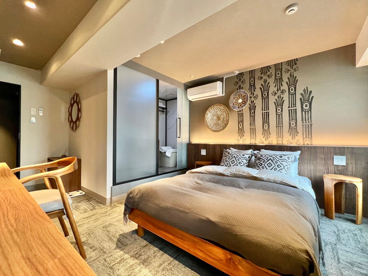 A well-appointed room features a spacious bed with a neatly arranged gray blanket. Decorative wall elements are present, enhancing the atmosphere. A small wooden table and chair are positioned nearby, while subtle lighting illuminates the soft textures of the carpet.